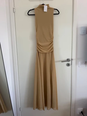 Beige/mörk senapsgul långklänning med lägre hals detalj - Säljer en elegant beige långklänning med hög hals och ärmlös design. Klänningen har en snygg rynkning i midjan som ger en smickrande siluett och ett mjukt, flytande fall. Perfekt för dig som gillar stilrena och minimalistiska plagg. Endast testad