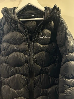 Svart dunjacka från Peak Performance - Snygg svart dunjacka från Peak Performance med vågigt quiltat mönster och dragkedja framtill. Jackan har huva och logga på bröstet. Perfekt för kalla höst/vår dagar  och riktigt lätt att bära. Klassisk design som funkar till det mesta. Använd en höst sen bara hängt i garderob och söker en ny ägare. Nypris 1899kr
