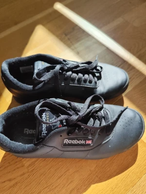 Svarta Reebok Classic sneakers - Snygga svarta Reebok Classic sneakers i läder med svart sula och svarta skosnören. Ikonisk logga på sidan och plös, samt diskreta perforeringar vid tårna för ventilation. Perfekta för en clean och stilren look.