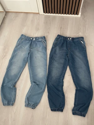 Två par baggy jeans med resår - Säljer två par baggy jeans med resår i midja och benslut. Ena paret är ljusblått och det andra är mörkare blått. Båda har fickor fram och knappstängning. Perfekta för en avslappnad och trendig stil. Blivit lite slitna i tvätten så sticker ut vit tråd ut från ett ”hål” på både byxorna