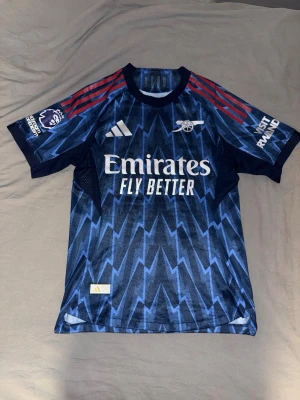 Arsenal blå bortatröja Adidas - Snygg Arsenal bortatröja i blått med blixtmönster, röda ränder på axlarna och vita loggor. Tröjan har Premier League-märke på ärmen och är tillverkad i ett lätt och ventilerande material. Perfekt för fotbollsträning eller match.