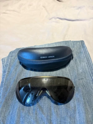 Svarta solglasögon Giorgio Armani - Snygga svarta solglasögon från Giorgio Armani med sportig, rundad sköldform och mörka linser. Bågarna är i svart plast och glasögonen kommer med originalfodral. Perfekta för dig som vill ha en stilren och modern look.