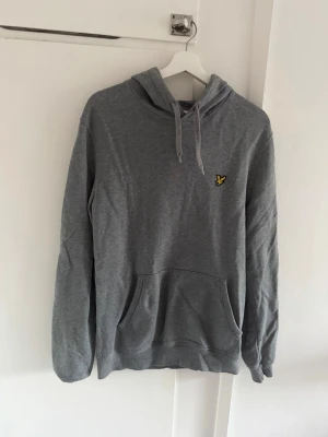 Grå Lyle & Scott hoodie  - Säljer en grå hoodie med huva och dragsnören samt en stor magficka. På bröstet finns en liten gul logga. Hoodien har långa ärmar och är gjord i mjukt material, perfekt för en avslappnad stil.
