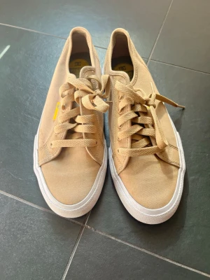 Beige sneakers med snörning - Snygga beige sneakers med klassisk låg profil och vit sula. Skorna har snörning och är tillverkade i ett slitstarkt tygmaterial. Perfekta för dig som gillar en clean och enkel stil med en touch av street.