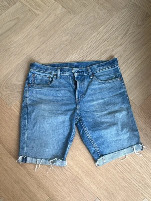 Blå Levis 511 jeansshorts med rå kant - Säljer ett par klassiska Levis 511 blå jeansshorts med rå, fransig kant och uppvik. Shortsen har fem fickor, bälteshällor och knappgylf. Ej klippa själv utan köpta som shorts. Perfekta för en avslappnad stil och tillverkade i slitstark denim. Storlek w31