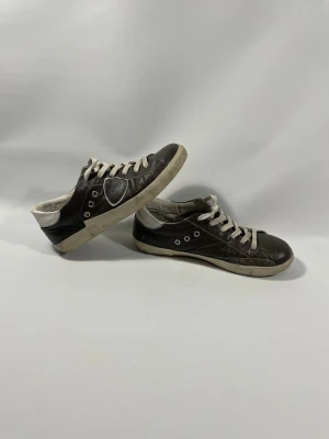 Philippe Model svarta sneakers - Bruna läder sneakers från Philippe Model med vita detaljer och vita skosnören. Skorna har en låg profil, rund tå och vit sula. Tillverkade i läder med märkets logga på hälen och sidan. Perfekta för en avslappnad och trendig stil. Jag tvättar dom rigält när jag skickar iväg dom 