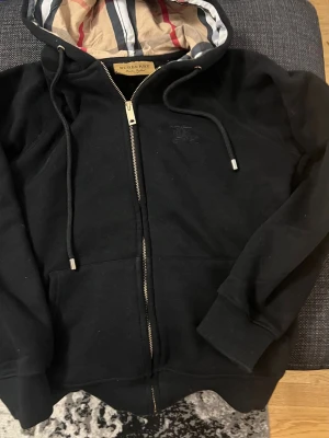 Svart zip hoodie från BB - Snygg svart hoodie från BB med dragkedja och klassiskt rutigt foder i huvan. Tröjan har dragsko i huvan, ribbade muddar och fickor framtill. Perfekt för en stilren och avslappnad look. Materialet är mjukt och bekvämt.