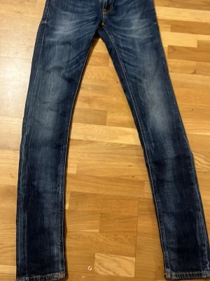 Blå slim fit jeans från Nudie - Snygga blå slim fit jeans från Nudie med klassisk femficksdesign och ljusa slitningar. Jeansen har guldfärgade sömmar och vågig brodyr på bakfickorna. Perfekta för en avslappnad och trendig look. Pris kan diskuteras.