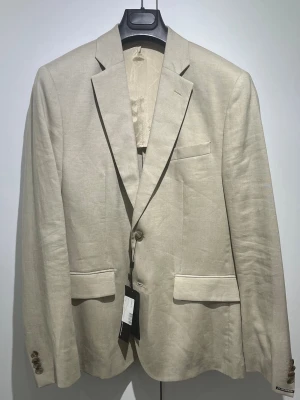 Beige linnékavaj - J.lindeberg - Stilren beige kavaj i linne med två knappar framtill och klassiska fickor med lock. Kavajen har smala slag och en bröstficka för en modern look. Perfekt för dig som gillar en avslappnad men ändå dressad stil.