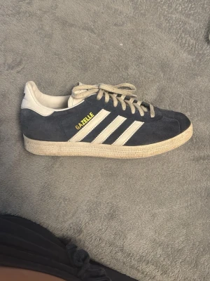 Adidas Gazelle mörkblå sneakers - Säljer ett par klassiska Adidas Gazelle sneakers i mörkblå mocka med vita detaljer och de ikoniska tre ränderna på sidan. Skorna har beige snörning och sula samt gul text på sidan. Perfekta för en sportig och stilren look.
