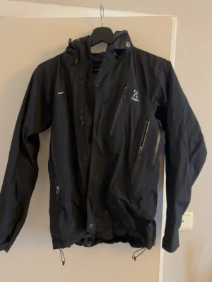 Svart vindjacka från Haglöfs - Snygg svart vindjacka från Haglöfs med flera praktiska dragkedjeförsedda fickor och justerbar huva. Jackan har en sportig look och är perfekt för blåsiga dagar. Tillverkad i slitstark syntet och har långa ärmar samt diskret logga på bröstet.