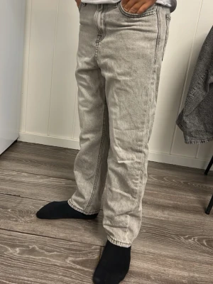 Grå raka jeans med tvättad look - Säljer ett par grå jeans med rak passform och tvättad finish. Jeansen har klassisk femficksdesign och synliga sömmar. Perfekta för en avslappnad och trendig stil. Materialet känns som bomullsdenim.