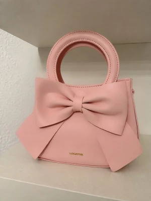 Rosa handväska med stor rosett - Supersöt rosa handväska med en stor dekorativ rosett på framsidan. Aldrig använd endast stått på hyllan. Väskan har rundade handtag och guldfärgade detaljer. Tillverkad i slätt konstläder och stängs med dragkedja. Medföljer även ett långt band. Perfekt för dig som gillar gulliga accessoarer.💛