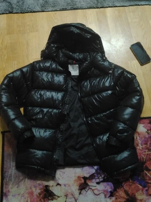 Moncler Maya Jacka  - Säljer en svart, glansig moncler jacka med huva och dragkedja. Jackan har breda, vadderade paneler och elastiska muddar vid ärmsluten. Perfekt för kalla dagar och riktigt snygg till streetwear-stilen. Har NFC tag