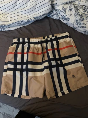 Beige-rutiga shorts med snörning - Snygga beige shorts med klassiskt rutigt mönster i svart, vitt och rött. Shortsen har elastisk midja med snörning och är perfekta för sommaren. Tillverkade i ett lätt syntetmaterial som känns skönt mot huden. Stilsäker detalj med logga på benet.