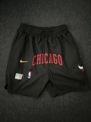 Chicago Bulls basketshorts svart - Snygga svarta basketshorts med Chicago Bulls-logga och texten CHICAGO i rött framtill. Shortsen har elastisk midja med dragsko, Nike-logga i gult och NBA-märke. Tillverkade i lätt och ventilerande material, perfekta för träning eller streetwear. Passar folk som är 160cm till 200cm. Den har hål i vänstra  fickan men ingenting märkes 