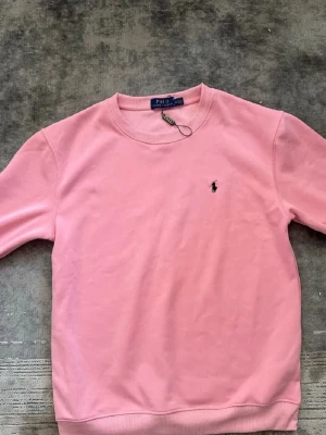 Rosa sweatshirt från Polo Ralph Lauren - Säljer en klassisk rosa sweatshirt från Polo Ralph Lauren med rund halsringning och broderad logga på bröstet. Tröjan har långa ärmar och ribbade muddar vid ärmslut och nederkant. Perfekt för en avslappnad och stilren look.