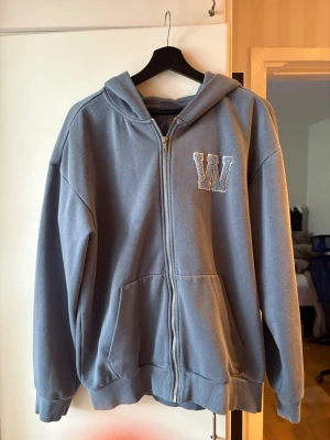 Blå hoodie med dragkedja och W-tryck - Säljer en blå hoodie med dragkedja och stor ficka framtill. På bröstet finns ett broderat W i vitt. Hoodien har huva och ribbade muddar vid ärmar och nederkant. Perfekt för en chill och avslappnad stil.