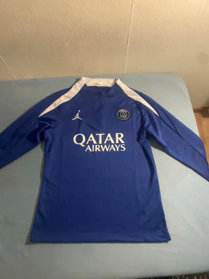 Blå PSG tracksuit - Säljer en blå långärmad träningströja från PSG med vita detaljer och halv dragkedja. Tröjan har tryck med Qatar Airways och Paris på ärmen samt klubbmärke och Jordan-logga på bröstet. Perfekt för fotbollsträning eller sportiga dagar.