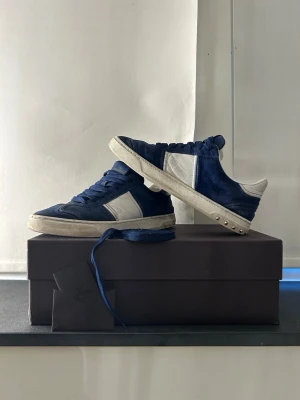 Valentino flycrew  - Snygga sneakers i blå mocka med vita detaljer och vita sidor. Skorna har blå skosnören och en vit sula med nitar på hälen. Låg modell som passar perfekt till streetwear och avslappnade outfits.