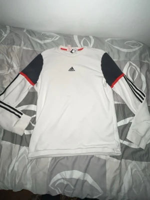 Vit långärmad tröja från Adidas - Vit långärmad tröja från Adidas med svarta paneler på axlarna och svarta ränder längs ärmarna. Tröjan har rund halsringning och röda detaljer vid axlarna. Perfekt för en sportig och avslappnad stil.