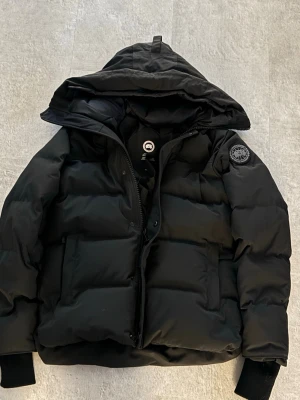  Canada Goose - Säljer en svart pufferjacka från Canada Goose med stor huva och klassisk patch på ärmen. Jackan har breda kanaler, två stora fickor framtill och muddar vid ärmsluten. Perfekt för kalla vinterdagar och riktigt snygg streetstil. Jackan är i storlek L och har en väldigt fin passform! 