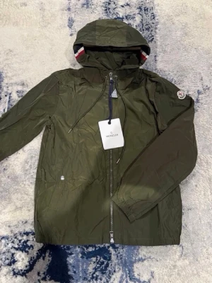 Grön windbreaker från Moncler - Stilren grön vindjacka från Moncler med huva och dragkedja framtill. Jackan har Moncler-logga på ärmen och en snygg detalj med rödvit rand på huvan. Tillverkad i lätt, vattenavvisande material och har två fickor med tryckknapp. Passar perfekt nu för hösten! Priset inte hugget i sten!🙌 passar M-L