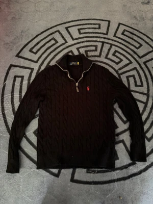 Svart kabelstickad tröja Polo Ralph Lauren - Kabelstickad svart tröja från Polo Ralph Lauren med halv dragkedja och hög krage. Klassisk röd logga broderad på bröstet. Tröjan har ribbade muddar och är tillverkad i ett mjukt, tjockt material som passar perfekt till kyligare dagar.