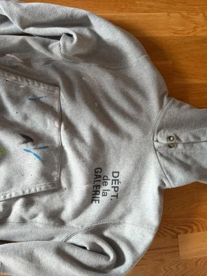 Grå hoodie DÉPT de la GALERIE - Säljer en grå hoodie från DÉPT de la GALERIE med tryck på framsidan och färgstänk över fickan. Hoodien har stor magficka, huva med knappar och är i mjukt bomullsmaterial. Perfekt för dig som gillar streetwear och unik design. Den är perfekt inför vintern då materialet är av väldigt högkvalite och väger mycket så man håller sig varm 