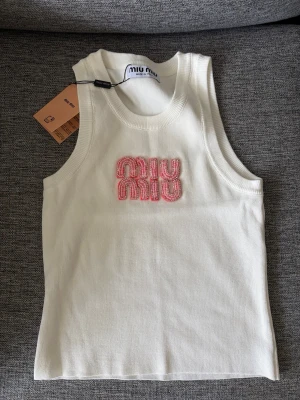 Vit ribbad linnetopp från Miu Miu - Säljer en vit ribbad linnetopp från Miu Miu med broderad logga i rosa och pärlor framtill. Toppen är ärmlös och har en klassisk rund halsringning. Perfekt för dig som vill ha en trendig och stilren topp med exklusiv känsla.