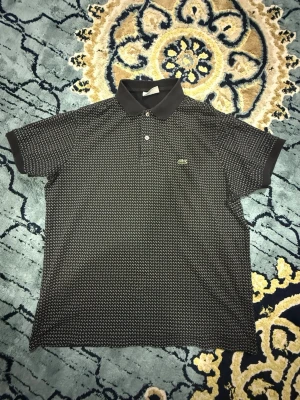 Svart mönstrad piké från Lacoste - Snygg svart pikétröja från Lacoste med vitt diskret mönster och klassisk krage. Tröjan har korta ärmar, två knappar framtill och den ikoniska gröna krokodilloggan på bröstet. Perfekt för en stilren och avslappnad look.