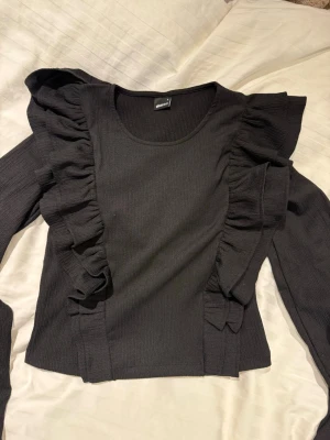 Svart topp med volanger - Snygg svart topp med långa ärmar och volangdetaljer längs axlar och framsida. Ribbat material som ger en skön passform och trendig look. Perfekt att styla med jeans eller kjol för en cool outfit.
