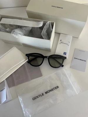 Svarta solglasögon från Gentle Monster - Snygga svarta solglasögon från Gentle Monster med klassisk rundad form och mörka linser. Bågarna är i blank svart plast och har diskreta detaljer vid gångjärnen. Perfekta för dig som vill ha en stilren och trendig look. Nypris 250€