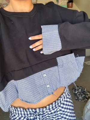 Svart croppad topp med rutig skjortdetalj - Unik croppad topp i svart med insydd rutig skjortdel i blått och vitt. Toppen har lång ärm och knappar framtill på skjortdelen. Perfekt för dig som gillar lager på lager och vill sticka ut lite extra.