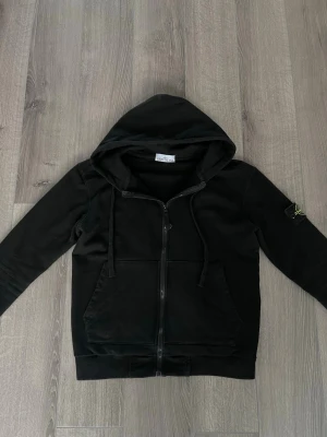 Stone island zip hoodie - Säljer en svart hoodie med dragkedja och huva. Hoodien har två stora fickor framtill och snörning i huvan. På vänster ärm sitter en patch med logga. Perfekt för dig som gillar en clean och stilren look.