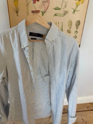 Randig skjorta från Polo Ralph Lauren - Klassisk vit skjorta med tunna blå ränder från Polo Ralph Lauren. Skjortan har button-down krage, långa ärmar och knappar framtill. Tillverkad i bomull och har en liten broderad logga på bröstet. Perfekt till jeans eller chinos.
