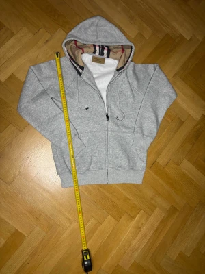 Grå hoodie med dragkedja och huva - ✅Storleken är S! Men passar som XS. Snygg grå hoodie med dragkedja och huva. Insidan av huvan har ett klassiskt rutigt mönster i beige, rött och svart. Tröjan har fickor framtill och ribbade muddar vid ärmslut och nederkant. Perfekt för en avslappnad och stilren look.