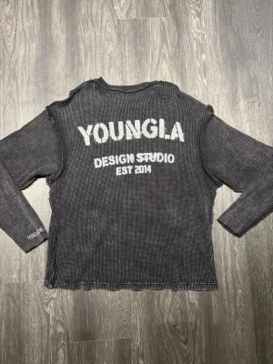 YoungLA Studio Long Sleeve svart/grå 2014 - YoungLA Design Studio Long Sleeve i svart/grå melerad design. Distinkt deconstructed look med synliga råa sömmar längs axlar, ärmar och sidor. Stort YOUNGLA DESIGN STUDIO EST. 2014-tryck på ryggen samt broderad YoungLA-detalj på ärmen. Figurnära atletisk passform med exklusiv streetwear-känsla. Exceptionellt fint skick, mycket välbevarad och nästintill som ny.