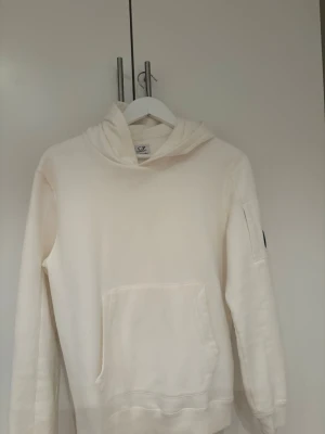 Vit hoodie från C.P. Company - Säljer en stilren vit hoodie från C.P. Company med klassisk huva och magficka. Ärmen har den ikoniska svarta linsen. Riktigt snyggt och knappt använd! Storlek 170 tveka inte att skriva om ni har nån fundering!