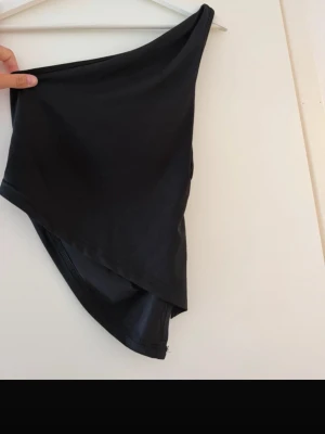 Svart oneshoulder tubtopp - Säljer en svart oneshoulder tubtopp med asymmetrisk skärning. Toppen är stilren och har en slät yta, perfekt för att skapa en edgy look. Materialet känns mjukt och följsamt mot huden.