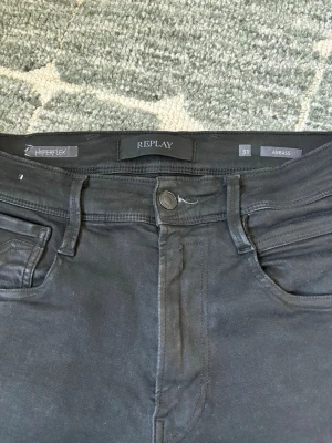 Svarta Replay Hyperflex jeans - Säljer ett par svarta Replay Hyperflex jeans i modellen Anbass. Jeansen har klassisk femficksdesign, smal passform och stretchigt material för extra komfort. Snygga detaljer som Replay-logga på bakfickan och patch bak i midjan.