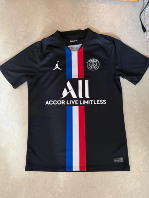 PSG x Jordan svart matchtröja - Snygg svart PSG matchtröja från Jordan med vertikala blå, vita och röda ränder framtill. Tröjan har klubbmärke på bröstet, Jordan-logga och sponsortryck i vitt. Tillverkad i lätt och ventilerande material, perfekt för fotboll eller träning.