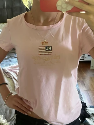 Ljusrosa RL t-shirt med tryck - Säljer en ljusrosa t-shirt med korta ärmar och ett snyggt RL-tryck framtill med krona och amerikansk flagga. T-shirten har en klassisk passform och är gjord i mjuk bomull, perfekt till jeans eller shorts.
