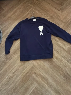 Mörkblå stickad tröja med hjärta - Mysig mörkblå stickad tröja med rund halsringning och ribbade muddar. På bröstet finns ett vitt hjärta och bokstaven A som detalj. Perfekt för dig som gillar enkel men snygg design. Passar till många olika stilar.