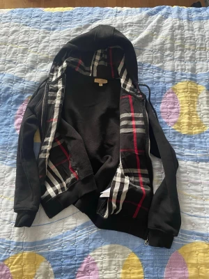 Svart hoodie med rutigt foder - Svart hoodie med dragkedja och huva. Insidan av huvan och öppningen har ett klassiskt rutigt mönster i svart, vitt och rött. Hoodien har fickor framtill och ribbade muddar vid ärmar och nederkant. Perfekt för en chill och stilren look.