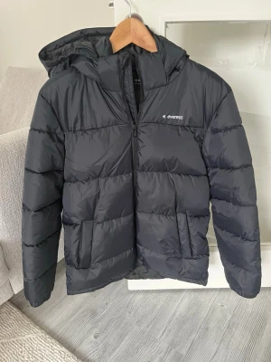 Svart dunjacka från Everest - Snygg svart dunjacka från Everest med huva och dragkedja framtill. Jackan har två stora fickor på sidorna och diskret logga på bröstet. Perfekt för kalla vinterdagar tack vare det tjocka dunfodret och den klassiska puffiga designen.
