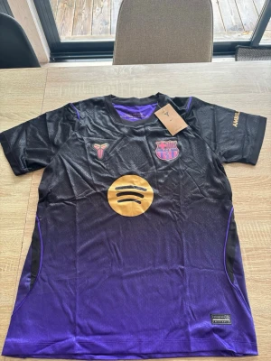 Svart-lila FC Barcelona Nike tröja - Snygg svart och lila FC Barcelona matchtröja från Nike med Dri-FIT-material. Tröjan har klubbmärke på bröstet, Kobe Bryant-logga, Spotify-logga i guld och lila detaljer längs sidorna. Perfekt för fotboll eller träning.