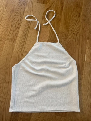 Vit croppad halterneck topp - Säljer en vit croppad halterneck topp med tunna knytband i nacken. Toppen har en enkel och stilren design med lätt struktur i tyget. Perfekt till sommaren och enkel att matcha med olika outfits.