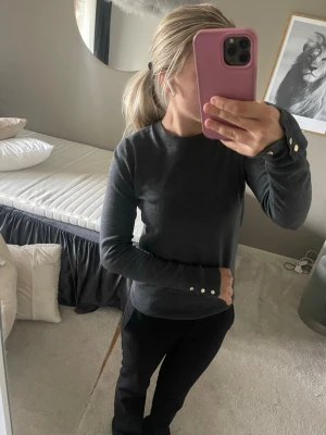 Mörkgrå stickad tröja med pärlknappar - Snygg mörkgrå stickad tröja med rund halsringning och långa ärmar. Tröjan har dekorativa pärlknappar vid ärmsluten som ger en extra detalj. Passar perfekt till både jeans och kostymbyxor. Materialet ser mjukt och bekvämt ut. Helt ny 