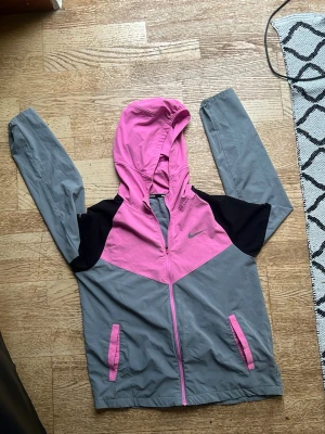 Nike vindjacka rosa och grå - Snygg vindjacka från Nike i rosa och grått med svarta detaljer. Jackan har huva, dragkedja framtill och två fickor med rosa dragkedjor. Tillverkad i lätt och funktionellt Dri-FIT-material, perfekt för löpning eller promenader.